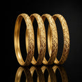 Four gold-toned bangles with detailed floral and geometric engravings, arranged in a row on a reflective black surface.