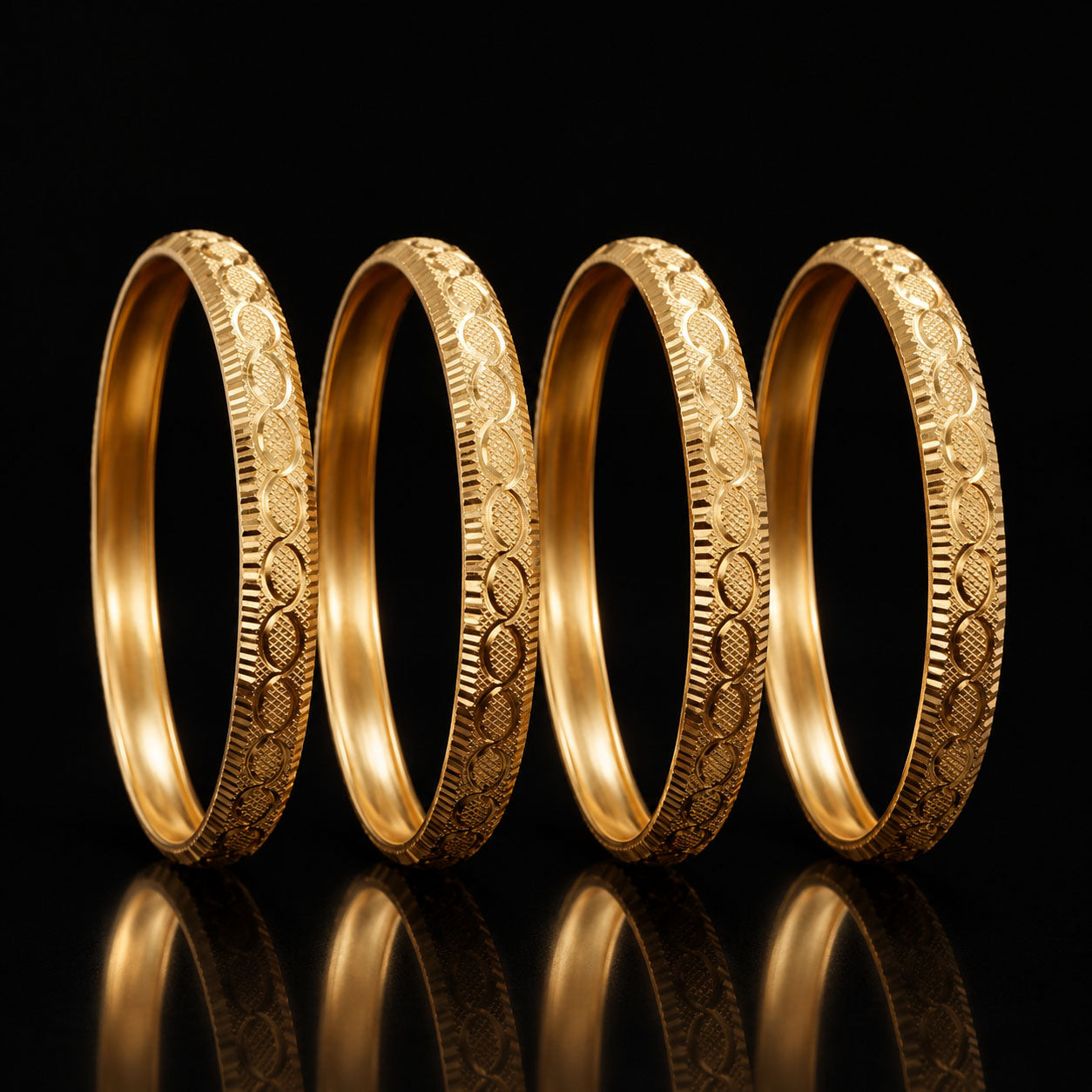 Four gold bangles with intricate floral and geometric engravings, arranged in a row against a solid black background with a reflective surface below.