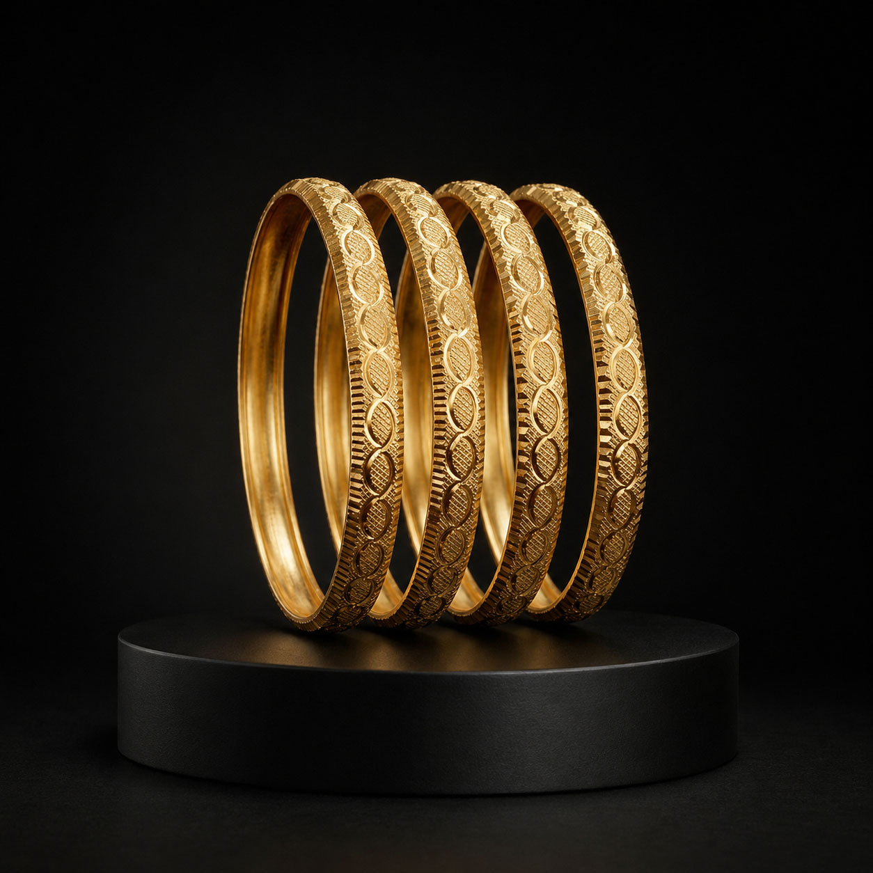Four gold bangles with intricate textured patterns, displayed on a black cylindrical pedestal against a dark background.