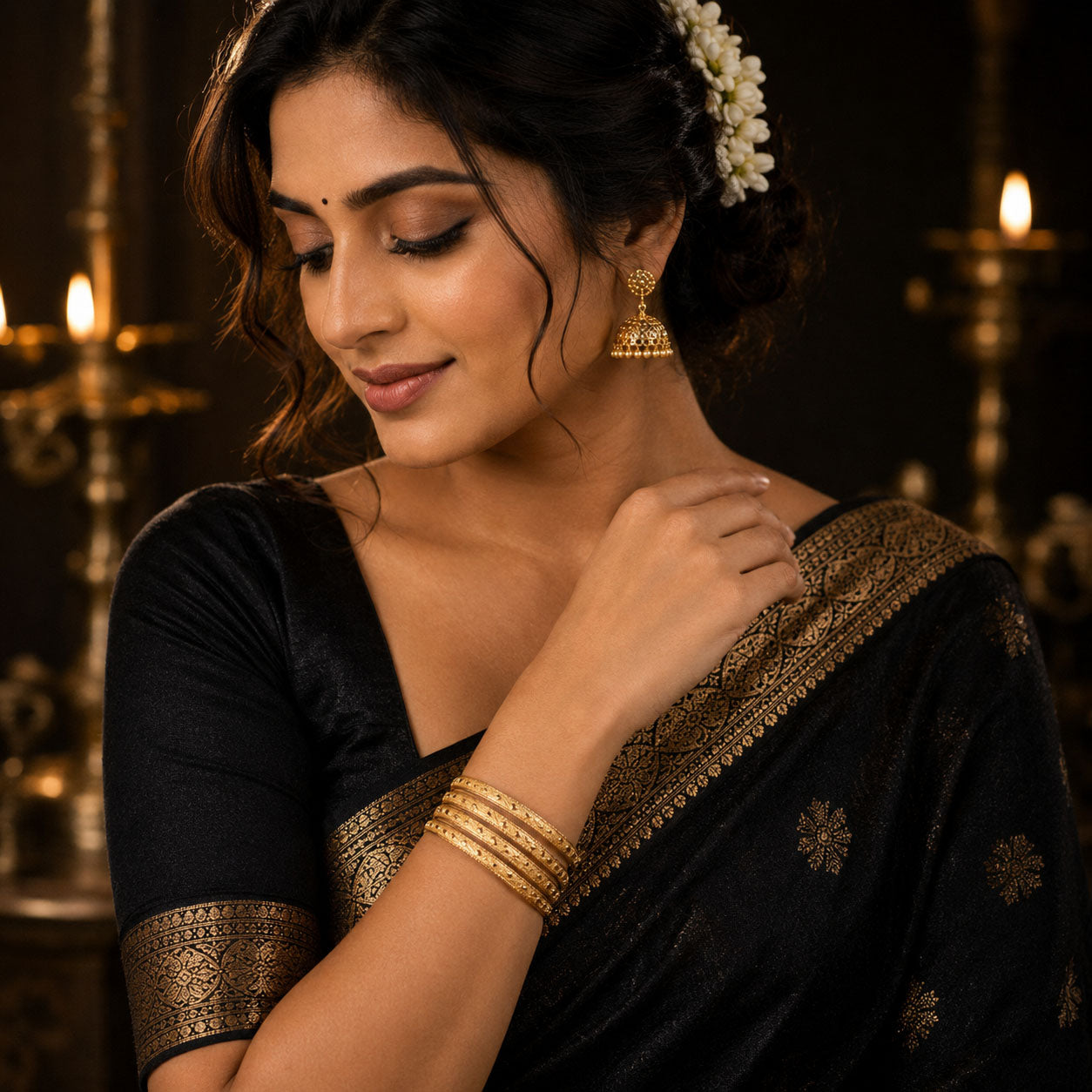 A woman wearing a black silk saree with gold zari border and floral motifs, paired with gold bangles and earrings, posed in front of a warm, candle-lit background.