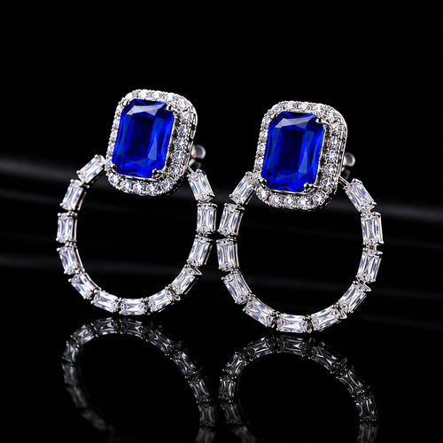 A pair of silver hoop earrings featuring a large rectangular royal blue gemstone in a halo setting, accented with baguette-cut clear stones, shown on a reflective black background.