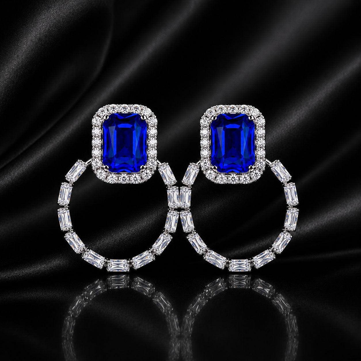 A pair of drop earrings featuring rectangular emerald-cut blue gemstones surrounded by small clear stones, hanging from silver-toned hoops set with baguette-cut clear stones, displayed on a black satin background.