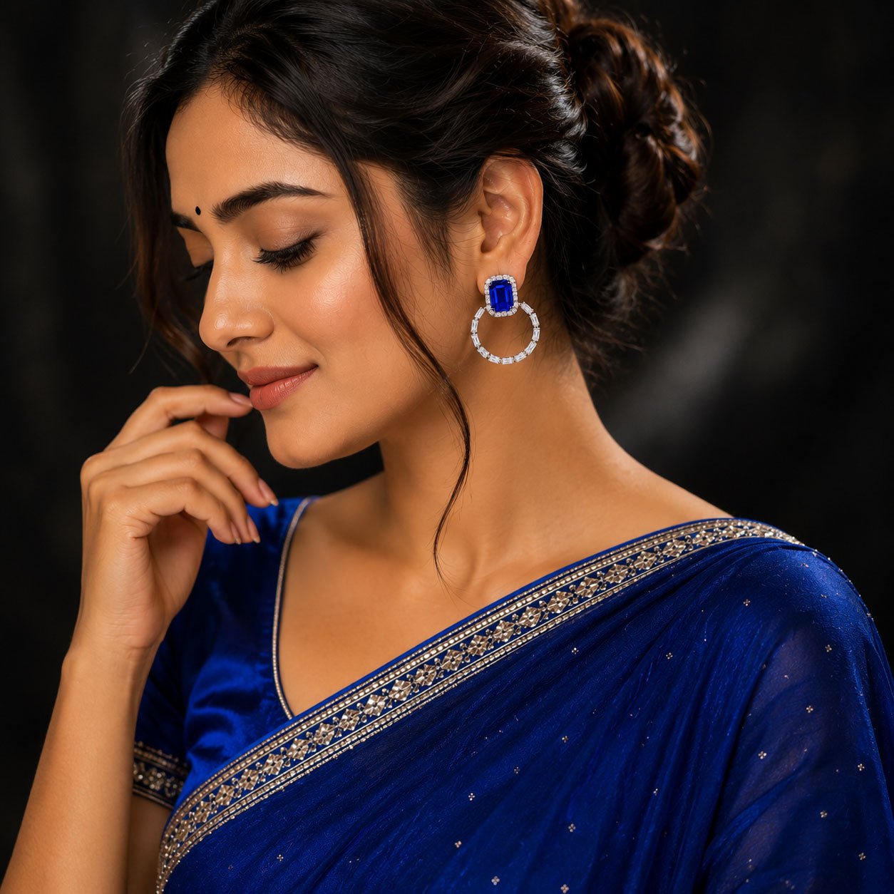 A woman wearing a royal blue saree with silver border embroidery and matching blue statement earrings with clear stones, posed against a dark background.