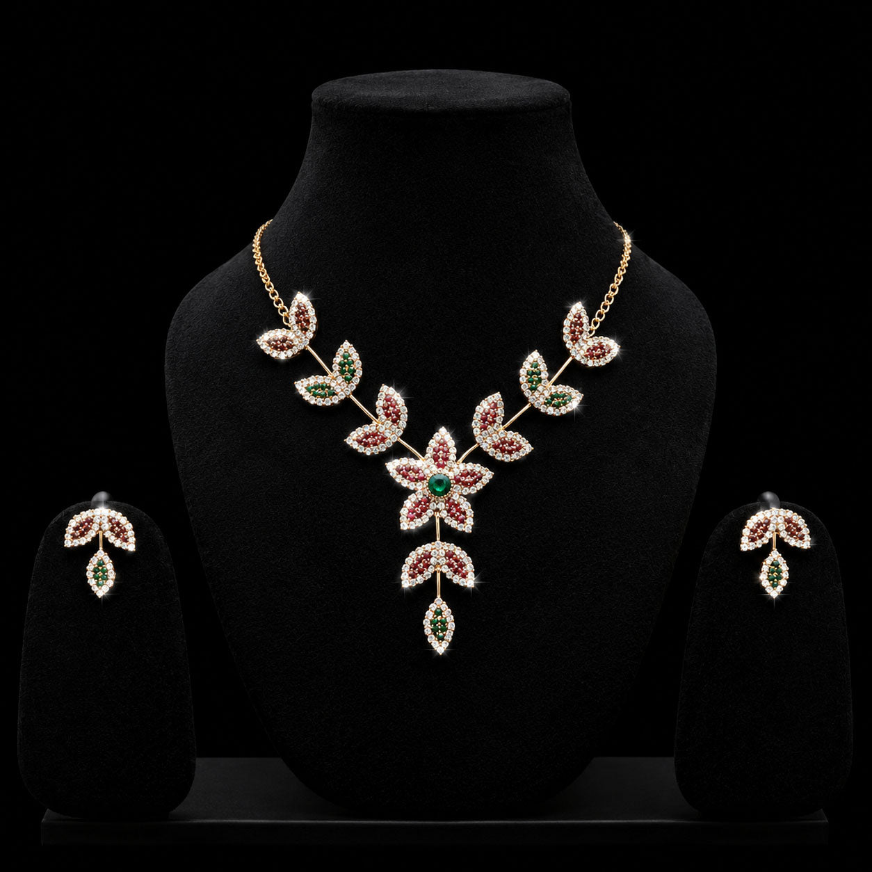 A gold-toned jewelry set displayed on a black velvet mannequin bust, featuring a leafy floral necklace with red and green stones, matching drop earrings, and a black background.