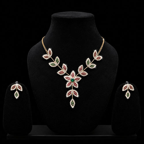 A gold-toned jewelry set displayed on a black velvet mannequin bust, featuring a leafy floral necklace with red and green stones, matching drop earrings, and a black background.