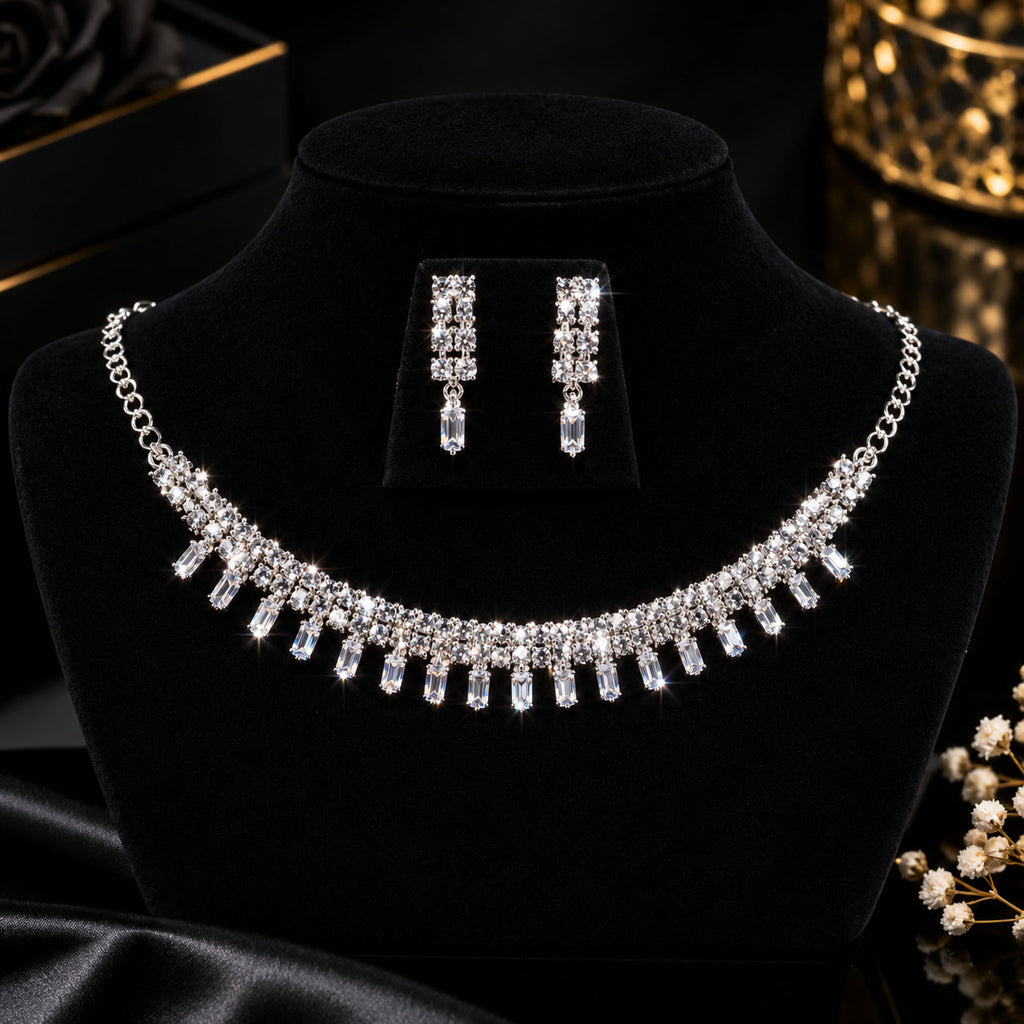 A silver-tone crystal necklace and matching drop earrings displayed on a black velvet jewelry bust. The necklace features a curved row of clear faceted crystals hanging from a silver chain, with the matching earrings shown on a small black card above it.