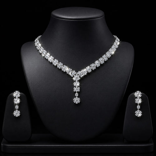A sparkling silver-tone jewelry set displayed on a black mannequin bust, featuring a V-necklace with a matching drop pendant and two matching drop earrings on small display stands against a black background.