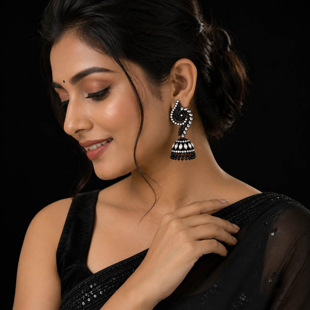 A woman wearing a black embellished saree and a matching black jhumka earring with silver-toned detailing, shown against a solid black background.