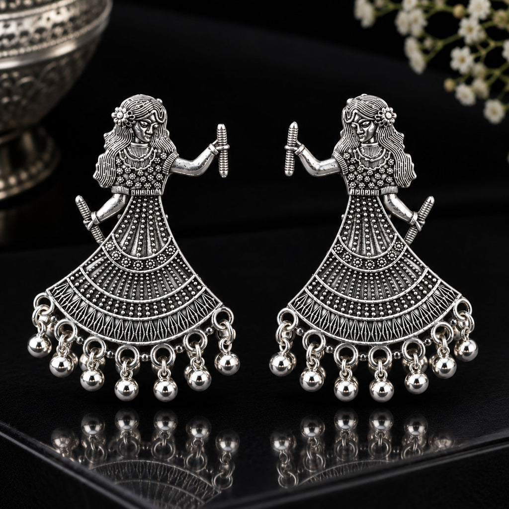 A pair of silver-toned traditional Indian Jhumka earrings featuring a dancing female figure design with intricate filigree work and dangling bell charms, displayed on a reflective black surface.