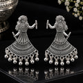 A pair of silver-toned traditional Indian Jhumka earrings featuring a dancing female figure design with intricate filigree work and dangling bell charms, displayed on a reflective black surface.