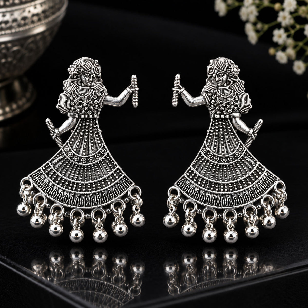 A pair of silver-toned traditional Indian Jhumka earrings featuring a dancing female figure design with intricate filigree work and dangling bell charms, displayed on a reflective black surface.