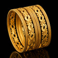 A stack of five wide gold bangles with intricate woven and floral patterns, displayed on a reflective black surface.