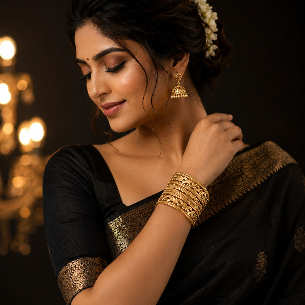 A woman wearing a black saree with gold border detailing and matching gold bangles, shown in a warm, dimly lit setting with blurred golden lights in the background.