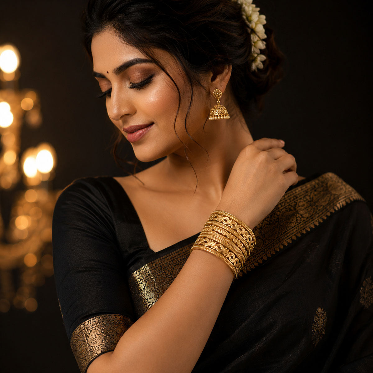 A woman wearing a black saree with gold border detailing and matching gold bangles, shown in a warm, dimly lit setting with blurred golden lights in the background.