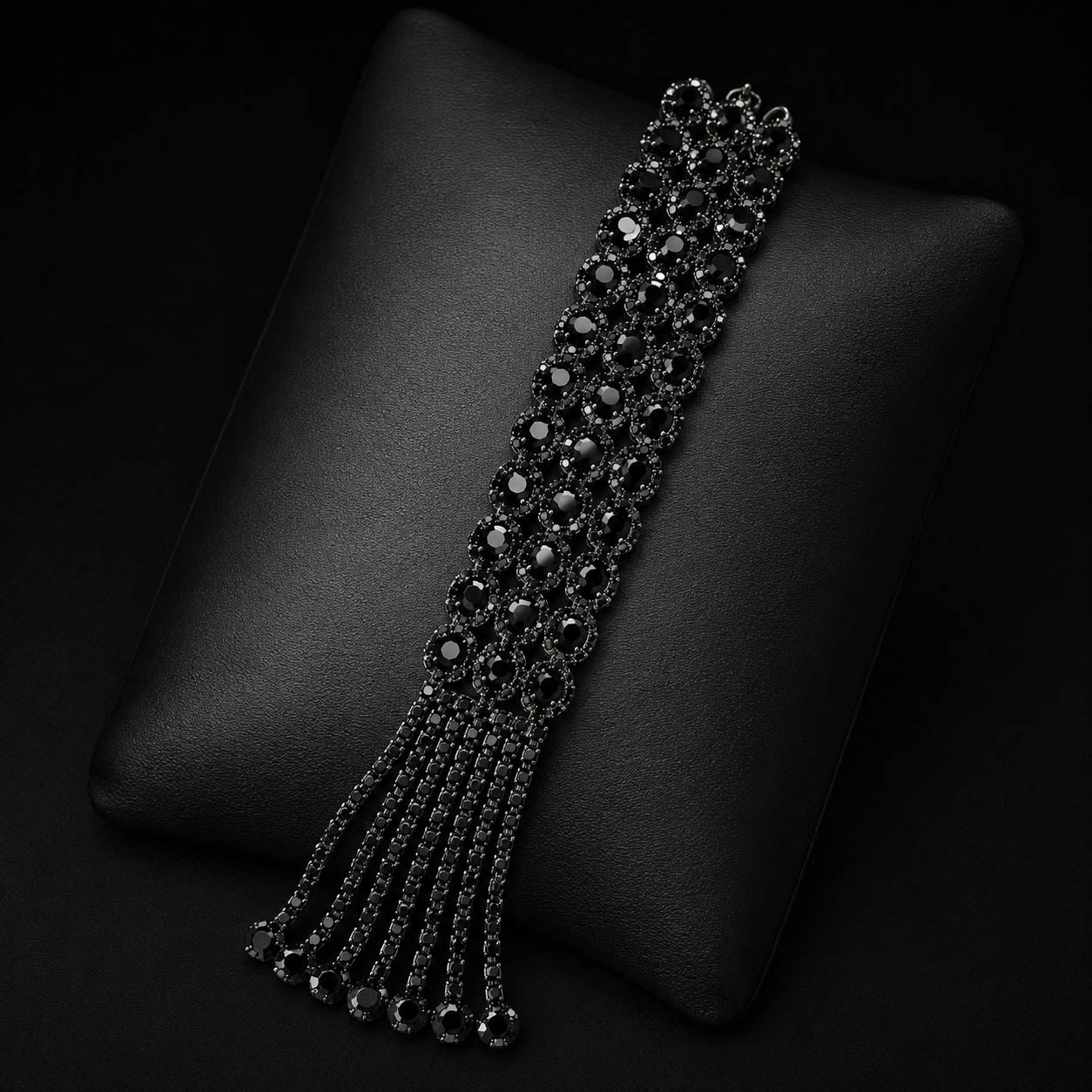 A black beaded tassel bracelet displayed on a black textured cushion against a dark background.