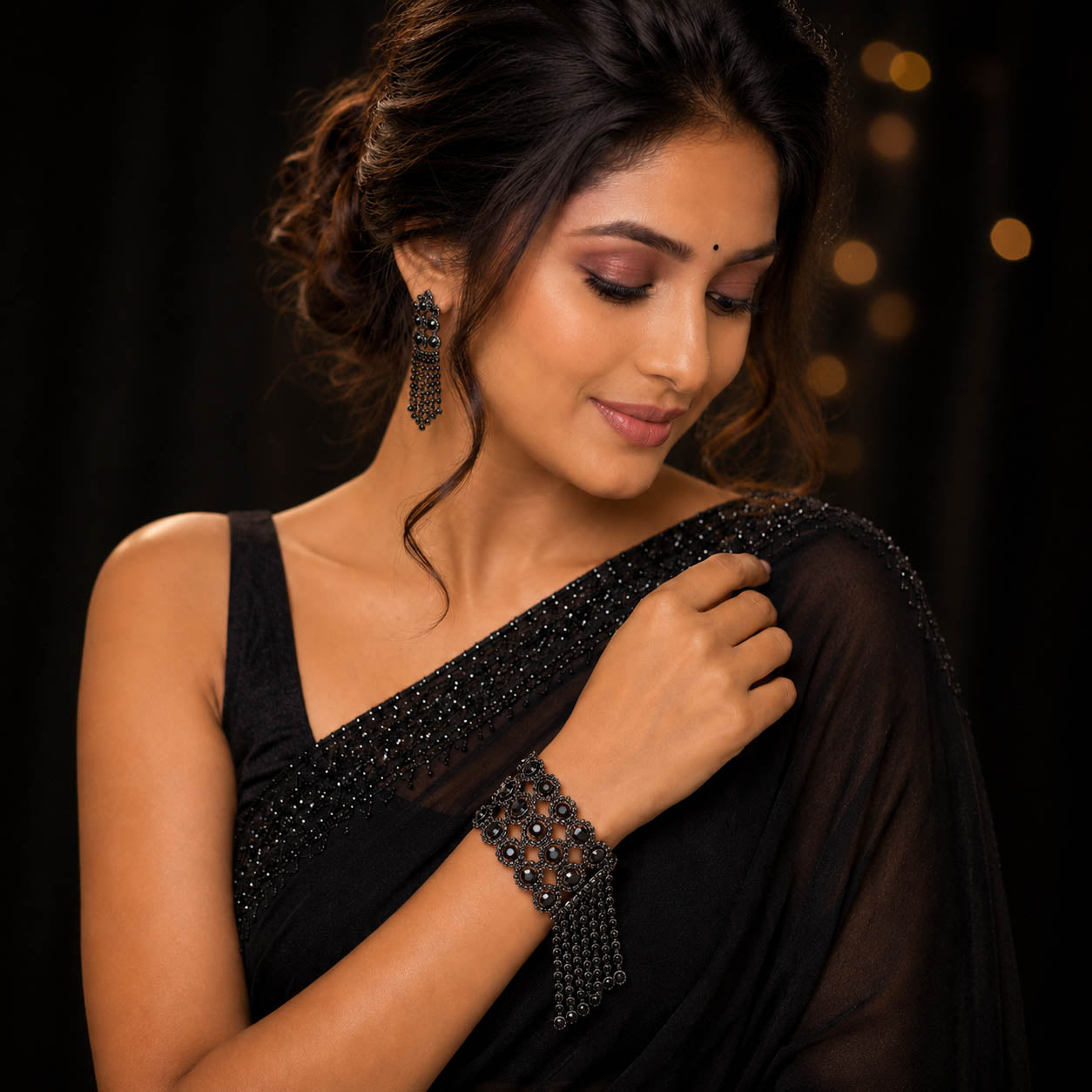A woman wearing a black embellished saree and matching black beaded jewelry, including a bracelet and earrings, posed against a dark background with soft warm bokeh lights.