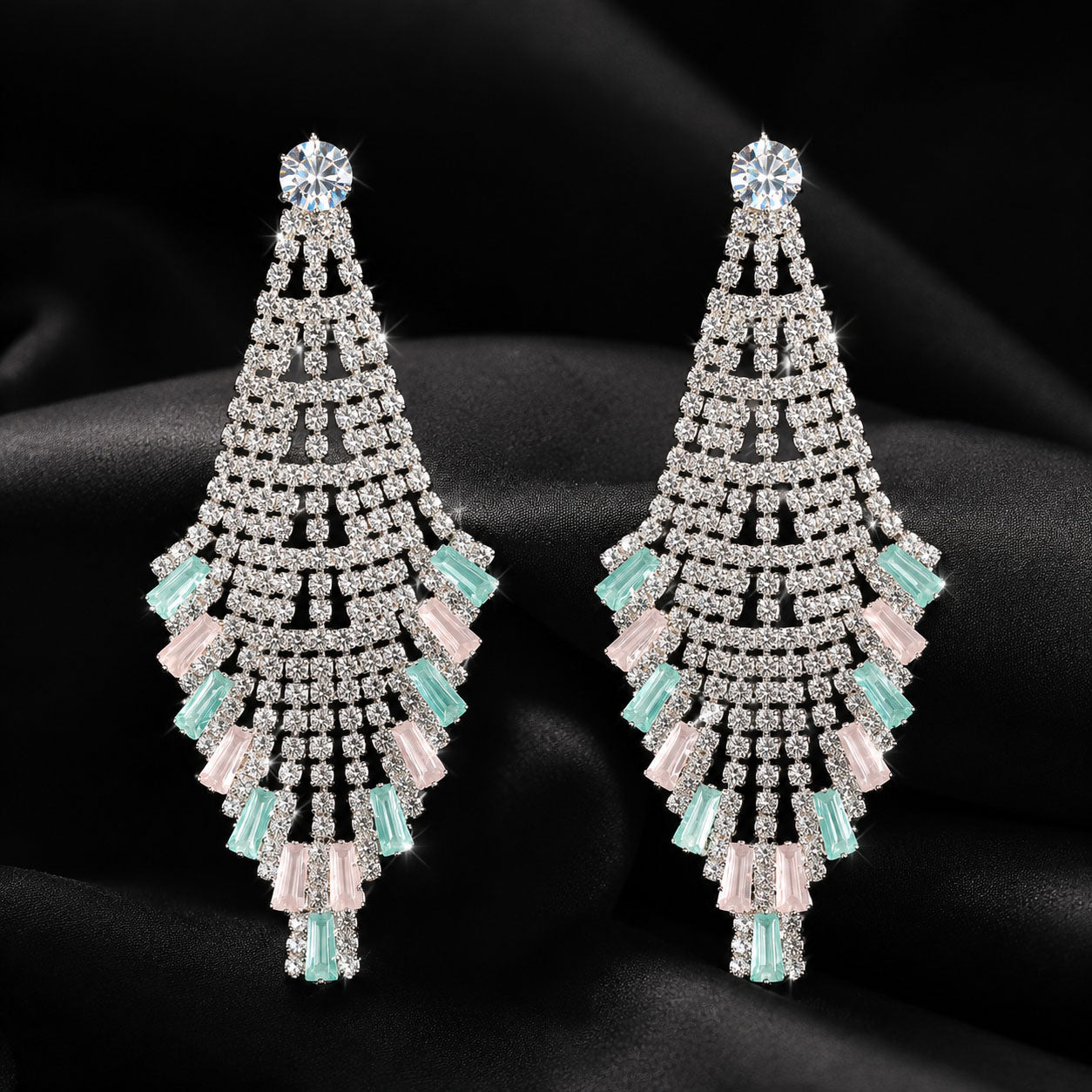A pair of large, dangling chandelier earrings with intricate silver-tone rhinestone detailing, accented by light pink and mint green crystals, displayed on a black satin fabric background.