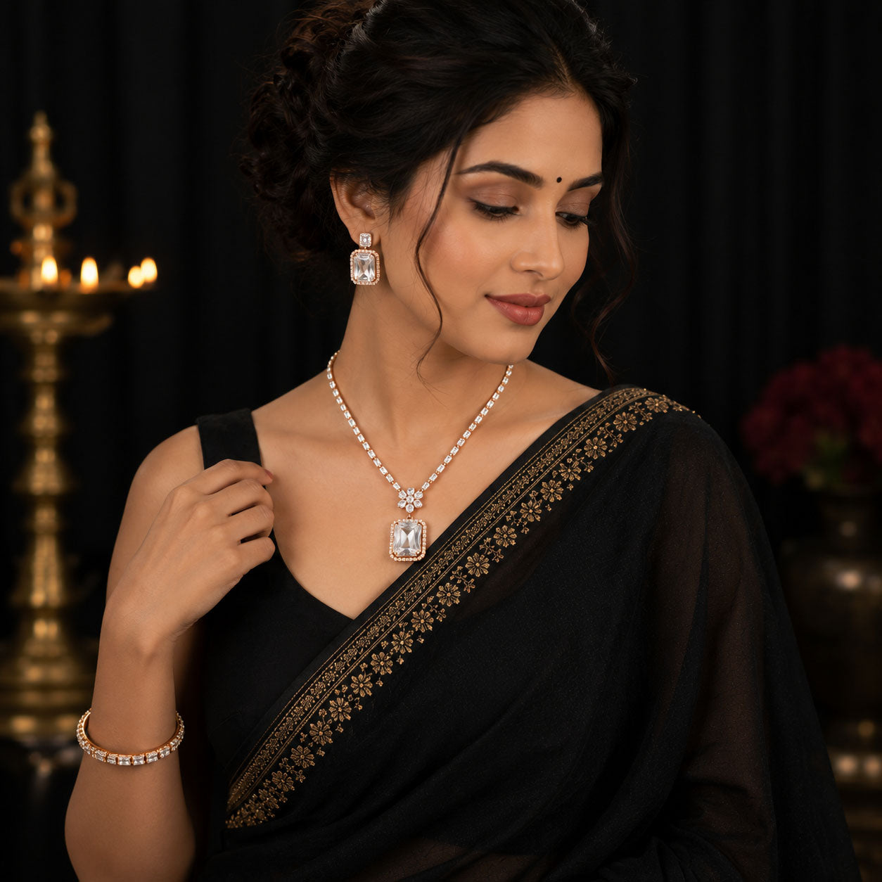 A woman wearing a black saree with gold border embroidery, styled with a pearl necklace featuring a rectangular clear gemstone pendant, matching drop earrings, and a pearl bracelet against a dark background with warm candlelight.