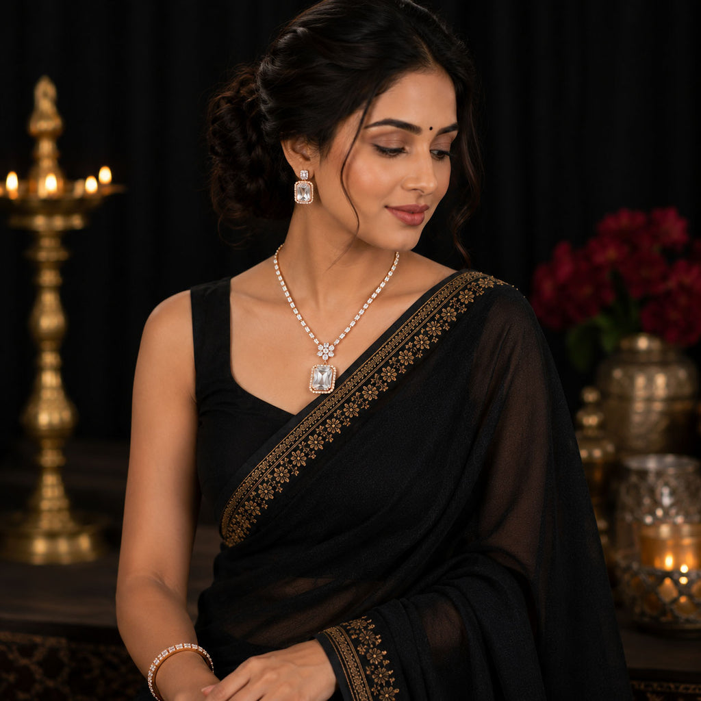 A woman wearing a black saree with gold border embroidery, styled with a pearl necklace, silver pendant, and matching earrings, posed in front of a dark background with warm decorative lights.