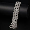 A silver-toned statement bracelet featuring a wide band of repeating floral clusters and a dangling fringe, displayed on a black cylindrical stand.