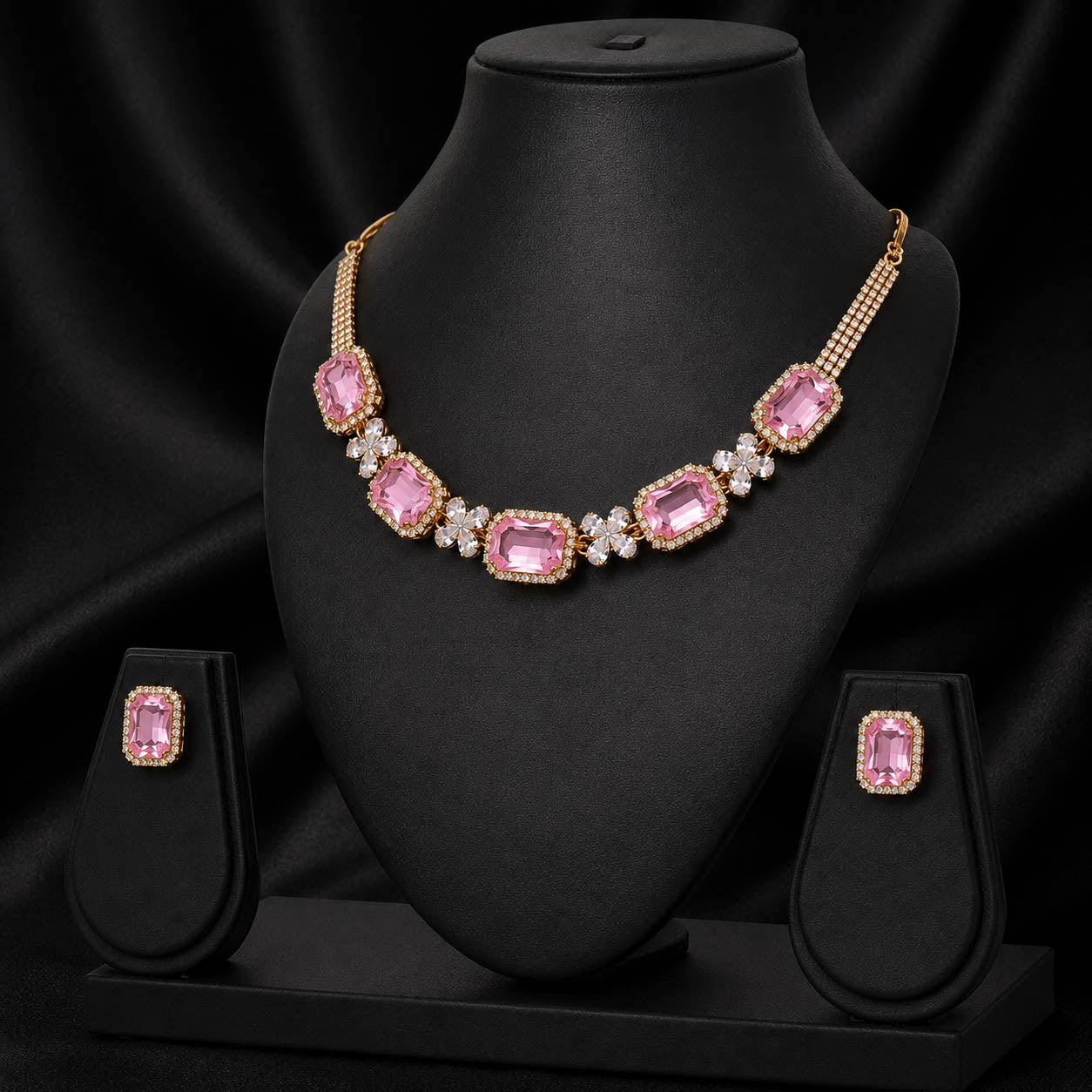 Gold-plated necklace and matching stud earrings with large rectangular pink gemstones and small clear accent stones, displayed on a black jewelry bust against a black satin background.