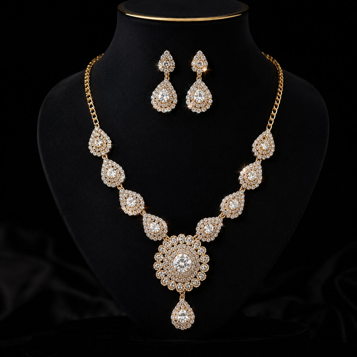 Gold-plated jewelry set featuring a statement necklace and matching teardrop earrings, both adorned with clear rhinestones, displayed on a black velvet mannequin bust.