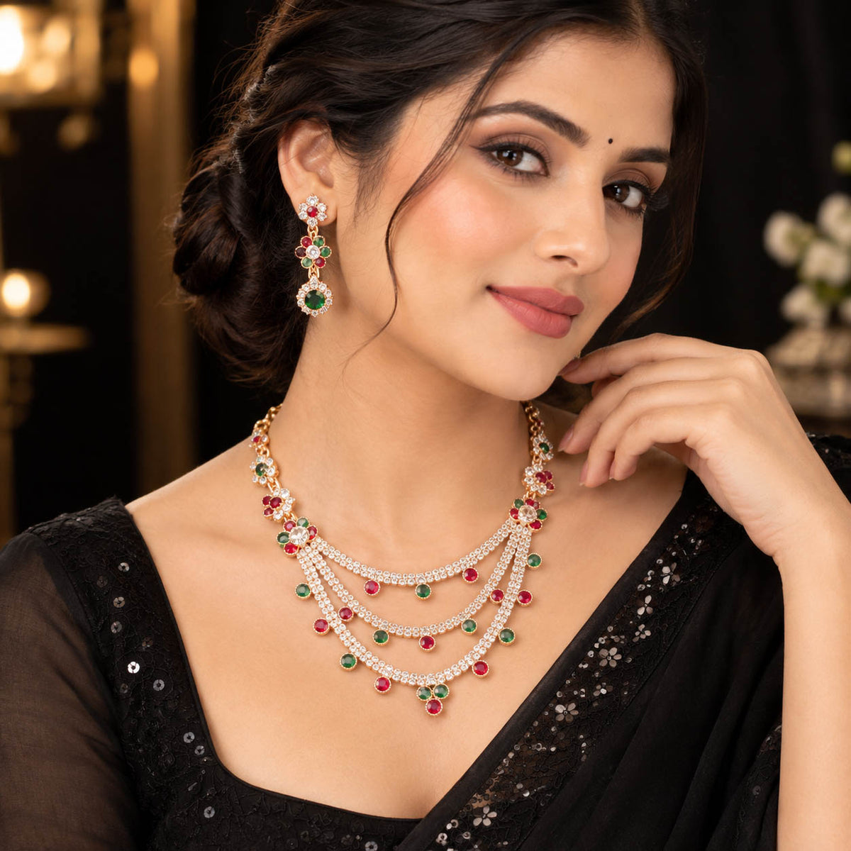 A woman wearing a multi-layered gold necklace with red and green gemstone accents, matching drop earrings, and a black embellished sari in a dimly lit indoor setting.