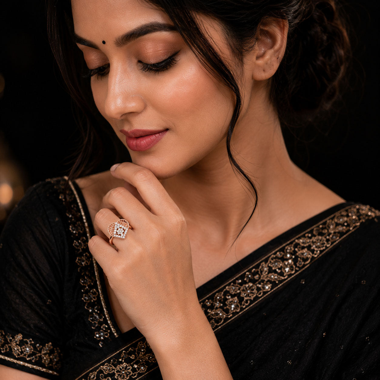 A woman wearing a black embroidered sari and a square diamond ring with a rose gold setting, posing with her hand near her chin against a dark background.