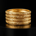 Four stacked gold bangles with intricate engraved patterns, displayed on a reflective black surface against a black background.