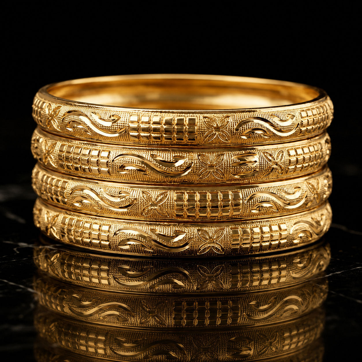 Four stacked gold bangles with intricate engraved patterns, displayed on a reflective black surface against a black background.