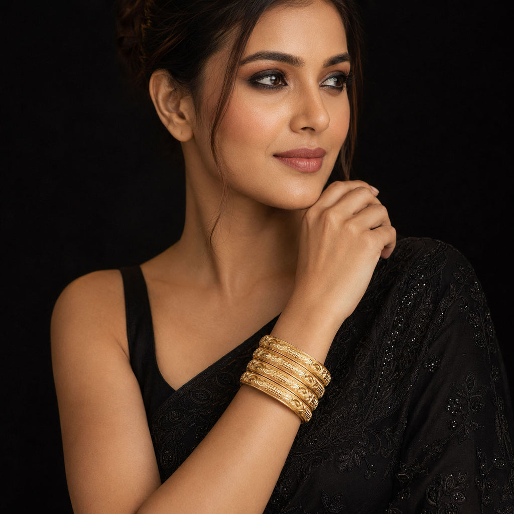 A woman wearing a stack of gold bangles on her wrist, posing in a black embroidered garment against a solid black background.