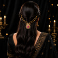 A woman shown from the back wearing a black traditional outfit with gold embroidery and a long, wavy black hairpiece adorned with a gold chain and a central red gemstone ornament. The background is a dark, elegant setting with blurred golden candlelight bokeh.
