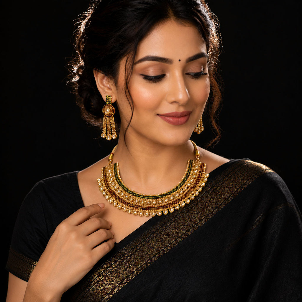 A woman wearing a black saree with a gold zari border, styled with a gold and green beaded necklace and matching gold earrings against a solid black background.