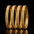 Four gold bangles with detailed geometric and chevron patterns, arranged in a row on a reflective black surface against a black background.
