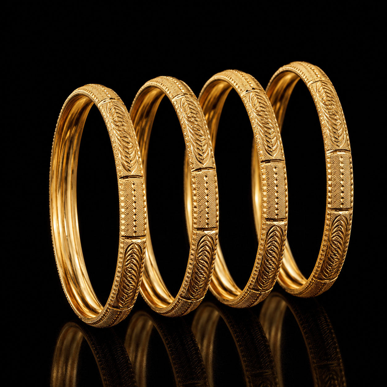 Four gold-toned bangles with detailed engraved patterns, arranged in a row against a solid black background with a reflective surface below.