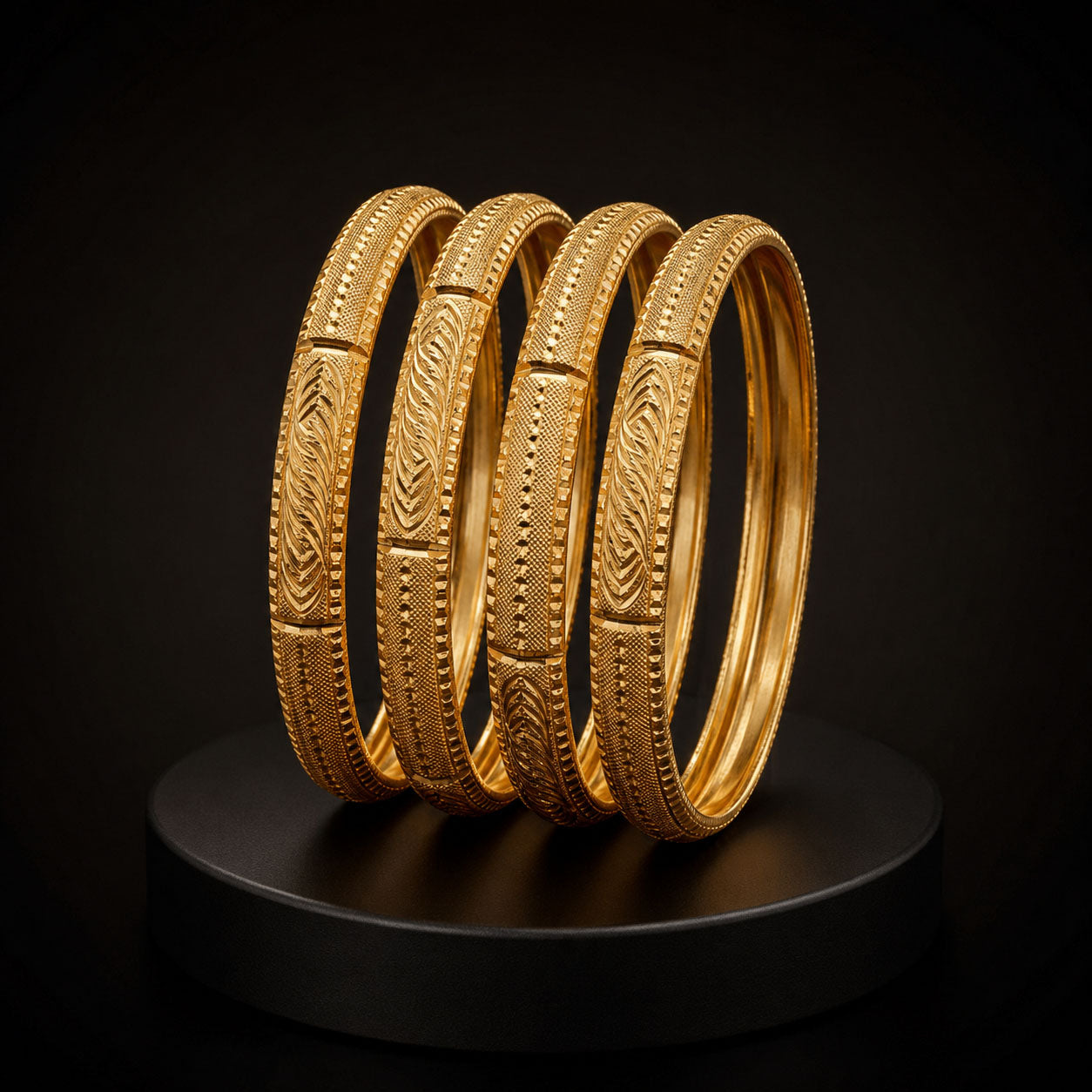 Four gold bangles with detailed engraved patterns, displayed on a black round pedestal against a dark background.