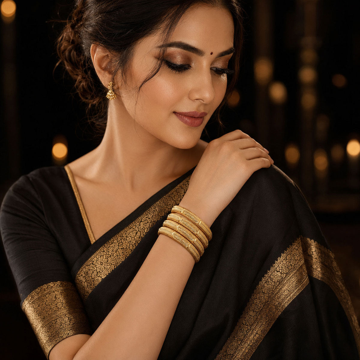 A woman wearing a black saree with a gold border and matching gold bangles, posed against a dark background with warm bokeh lights.