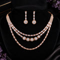A rose gold jewelry set displayed on a black velvet bust, featuring a three-layer necklace with baguette and round crystal settings, matching drop earrings, and a black box in the background.