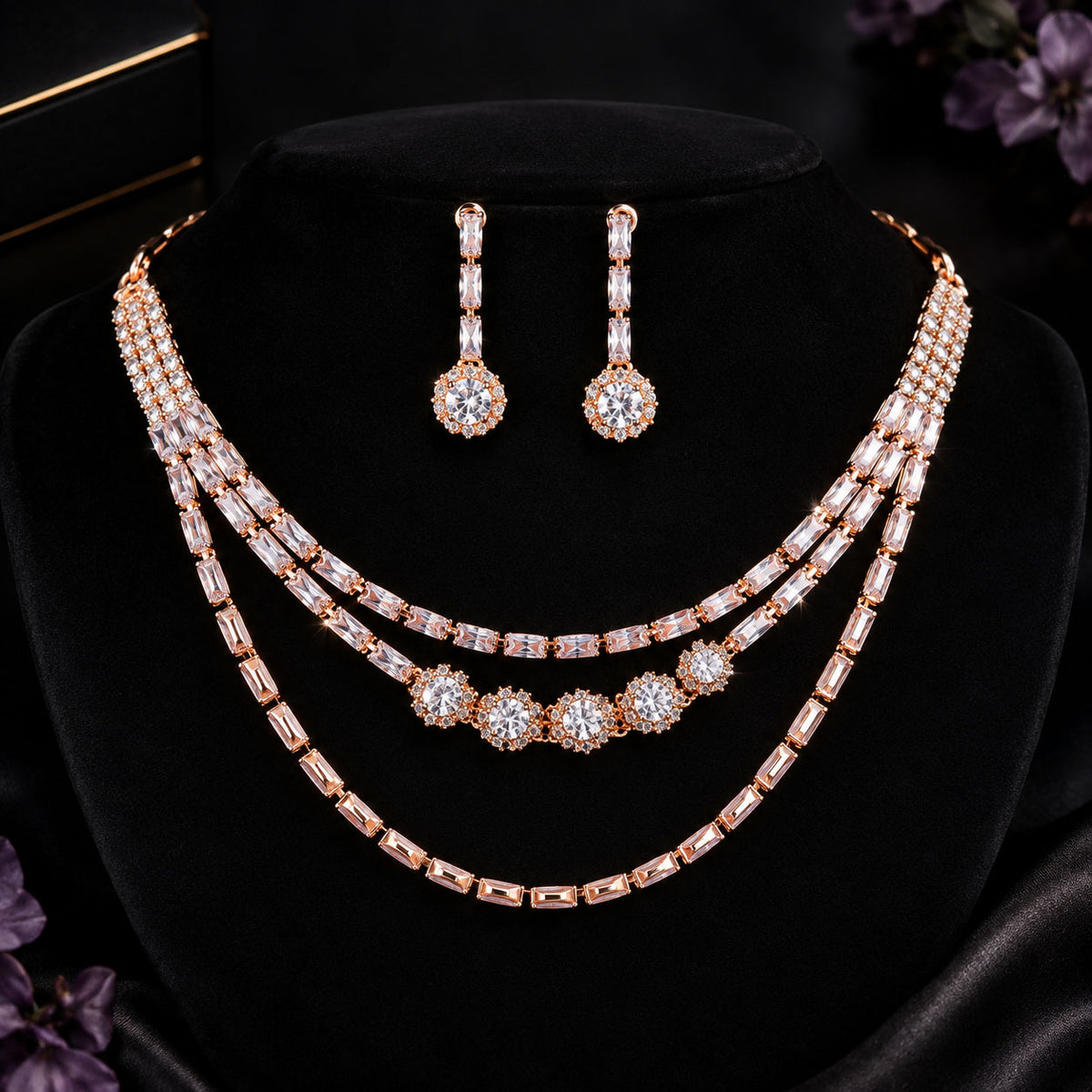 A rose gold jewelry set displayed on a black velvet bust, featuring a three-layer necklace with baguette and round crystal settings, matching drop earrings, and a black box in the background.